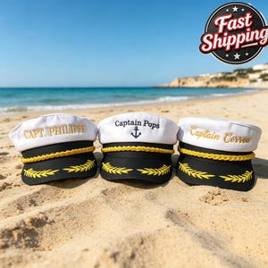 Könnte beinhalten: Drei weiß-schwarze Kapitänsmützen mit goldener Kordelverzierung und gold gestickten Lorbeerblättern auf dem Schirm. Die Mützen tragen individuelle Stickereien: Capt. Philippe, Captain Pops mit Anker-Symbol und Captain Correa in Gold oder Schwarz.