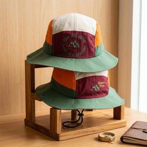 Könnte beinhalten: Ein farblich abgesetzter Fischerhut mit Einsätzen in Creme, Orange, Dunkelrot und Grün. Die Vorderseite zeigt eine grüne Stickerei mit dem Text Not All Who Lost Are Wander und einem Bergsymbol. Darunter ein zweiter Hut mit Roam Free Text.