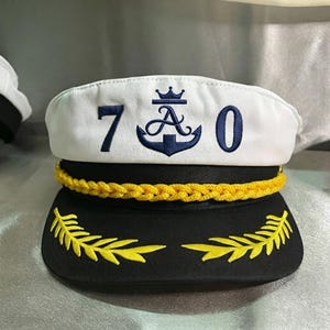 Op de afbeelding: Een wit met zwarte kapiteinspet met een donkerblauw geborduurd ankerlogo, een kroon en het getal 70. De pet is voorzien van een geel gevlochten koord aan de voorzijde en geel geborduurde laurierbladmotieven op de zwarte klep van de pet.