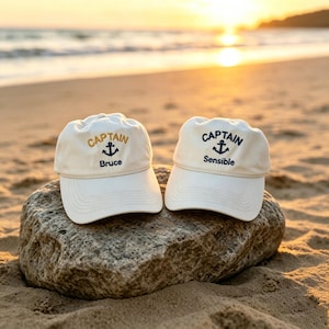 May include: Two off-white baseball caps featuring embroidered text and an anchor icon. The left cap displays the word CAPTAIN in gold and Bruce in navy blue. The right cap displays the word CAPTAIN and Sensible in navy blue text.