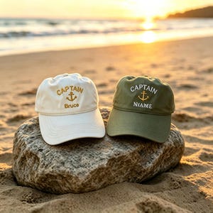 Könnte beinhalten: Zwei Baseballkappen, eine cremeweiß und eine olivgrün, jeweils mit einem gestickten goldenen Ankersymbol über dem Wort CAPTAIN und einem personalisierten Namen in weißem oder goldenem Garn, platziert auf einem großen Felsen am Strand.