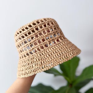 Puede incluir: Un sombrero de pescador de color tostado claro hecho de un material tejido y texturizado. El sombrero presenta un patrón de ganchillo abierto tipo rejilla en toda la copa y un ala sólida y tejida apretada que se extiende desde la base.