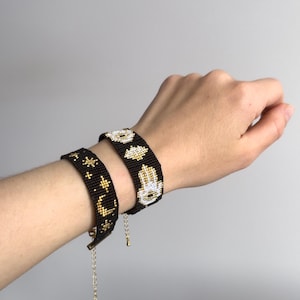 May include: Two black beaded bracelets featuring gold and white seed bead patterns. One bracelet displays a crescent moon and star motifs, while the other showcases a hamsa hand symbol. Both bracelets include gold-toned metal chain extenders.