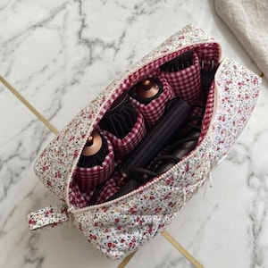 May include: Floral print cosmetic bag with a red and white gingham interior. The bag is open, revealing hair styling tools, including round brushes and a curling iron. The bag has a zipper closure and a small handle.