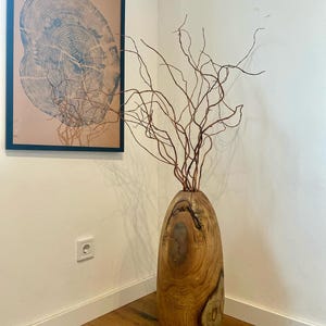 May include: A tall, cylindrical wooden vase with a natural grain pattern and a smooth, polished finish. The vase contains a cluster of thin, bare, twisted brown branches that extend upwards, creating a complex, branching silhouette.