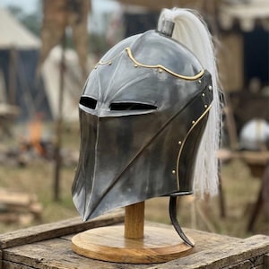 Puede incluir: Un casco de estilo medieval hecho de metal oscuro, con visera y ribete dorado decorativo. El casco tiene una pluma blanca y se exhibe sobre un soporte de madera, sobre un fondo borroso al aire libre.