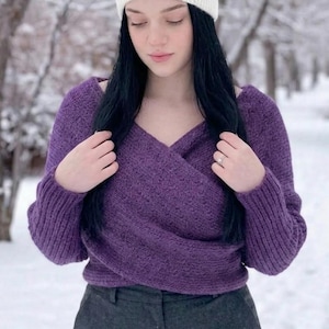 Op de afbeelding: Een persoon draagt een paarse wikkel trui en een grijze broek, gecombineerd met een witte gebreide hoofdband. De trui heeft een V-hals. De achtergrond is een besneeuwde buitenscène.