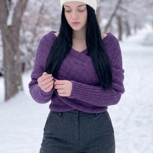 May include: A woman wearing a white knit headband, a purple sweater, and gray pants stands in a snowy outdoor setting. The sweater has a V-neck design. The background features snow-covered trees.