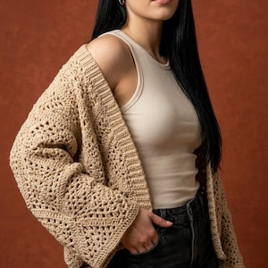 Könnte beinhalten: Beigefarbene Häkeljacke mit offenem Schnitt und strukturiertem Muster. Sie wird über einem cremefarbenen Rippen-Tanktop und dunkelgrauen Jeans getragen. Der Hintergrund ist ein warmer, brauner Farbton.