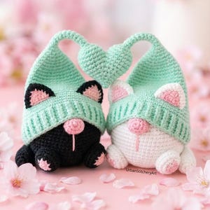 Pode incluir: Dois gnomos de gato de crochê feitos à mão, um preto e outro branco, usando chapéus verde-menta com detalhes em forma de coração. Os gnomos têm narizes e orelhas cor-de-rosa, em um fundo rosa com pétalas de flores.