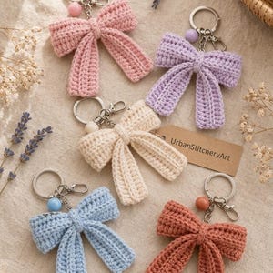 May include: Five crochet bow keychains in pink, lavender, cream, light blue, and terracotta. Each bow features a metal keyring, a lobster clasp, and a small round bead. A brown paper tag with the text UrbanStitcheryArt is attached to the cream bow.