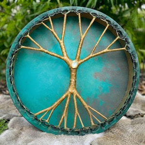 Pode incluir: Um tambor de moldura circular com uma pele turquesa e verde-azulado vibrante com acabamento manchado. Um design de árvore dourada com ramos ramificados está centrado na pele, acentuado por uma pequena pedra branca iridescente no centro.