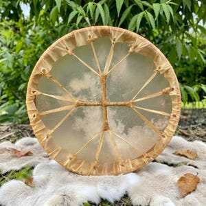 May include: A circular frame drum featuring a translucent, light-colored animal hide stretched tightly across a wooden hoop. The drumhead is secured with thin, light brown leather lacing that forms a cross pattern in the center.