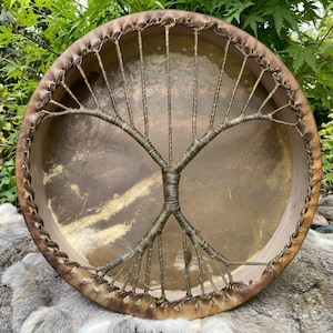 May include: A round frame drum with a stretched tan animal hide head. The drum features a decorative tree design created with dark brown cordage wrapped around the tension strings, which are secured to the circular wooden frame rim.