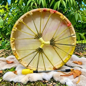 May include: A round frame drum with a light-colored, translucent skin head featuring yellow and red hues. The drum is laced with thin, light-colored cords that meet at a central point. A matching drum beater with a yellow head is included.