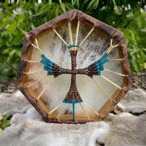May include: A hexagonal frame drum with a light-colored animal hide head. The frame is made of dark brown wood. The back features a cross-shaped handle woven with dark brown and teal cord, connected to the frame by light-colored tension cords.