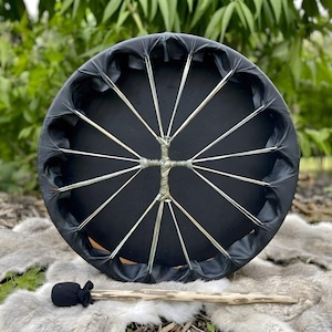 May include: A circular frame drum with a solid black drumhead and a black rim. The drum features a complex lacing system made of thin, light-colored cords that converge at a central point. A wooden drum mallet with a black fabric head is included.