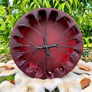 May include: A circular frame drum featuring a deep red drumhead surface. The drumhead is secured to the frame by dark brown leather lacing that radiates from a central point, creating a web-like pattern across the back of the instrument.