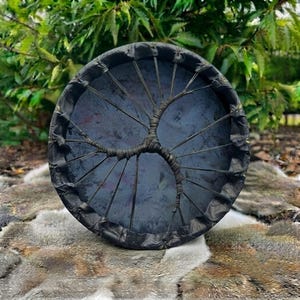 Op de afbeelding: Een ronde frametrommel met een donker, gemarmerd oppervlak. De trommel is voorzien van een zwarte, lederachtige rand en een centraal, boomvormig veterpatroon van dun, donker koord dat het trommelvel aan het frame bevestigt.