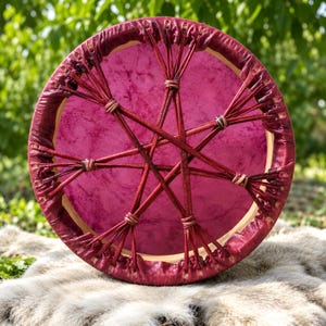 Pode incluir: Um tambor de moldura circular com uma pele de tambor magenta vibrante. O tambor é construído com um aro de madeira clara e é firmemente atado com tiras de couro vermelho escuro que formam um padrão de estrela na superfície frontal.