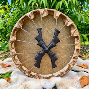 May include: A circular frame drum with a tan drumhead, featuring a tan leather rim and a central handle formed by two intersecting black leather-wrapped grips. The drum is tensioned by thin cords radiating from the center to the outer edge.