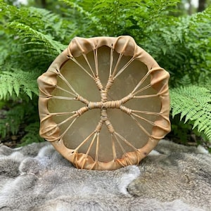 May include: A circular frame drum featuring a translucent, light-colored drumhead stretched over a wooden frame. The drumhead is secured by a network of thin, light brown leather lacing that radiates from a central point, creating a star-like pattern.