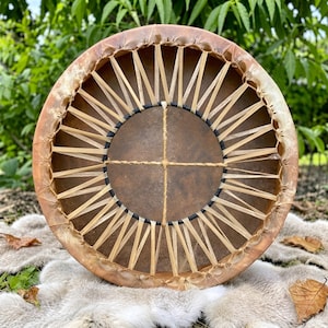 May include: A circular frame drum featuring a stretched tan animal hide head secured by a complex lacing system of thin, light-colored strips. The center of the drum head is marked by a cross-shaped lacing pattern, creating a geometric design.