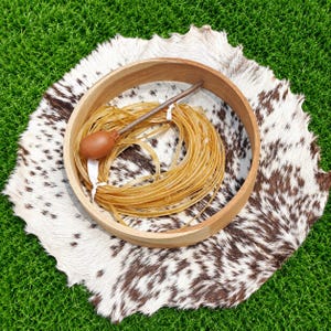 May include: A round wooden frame containing a coiled bundle of thin, translucent, light-tan plastic tubing. A single, smooth, brown, oval-shaped object is attached to the end of a thin, straight, brown stick resting atop the coiled tubing.