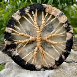 May include: A circular frame drum featuring a natural hide drumhead and a rim covered in black and tan animal fur. The back of the drum is laced with light tan cordage arranged in a cross pattern, secured with tight, intricate woven bindings.