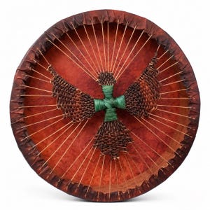 May include: A circular drum with a deep reddish-brown skin stretched over a wooden frame. Thin light-colored cords radiate from the center to the rim. A woven dark brown bird-shaped figure with a green wrapped center is attached to the cords.