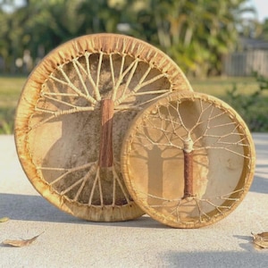 May include: Two handcrafted, round drums with a tree-of-life design. The larger drum is approximately 18 inches in diameter, and the smaller drum is about 10 inches. Both drums feature a natural, light brown hide stretched over a wooden frame, with a tree-like pattern.