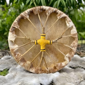 Puede incluir: Un tambor de marco circular con una piel de animal de color bronceado claro y borde de pelaje en el exterior. El tambor está tensado con finos cordones de cuero que convergen en el centro, envuelto en cuero amarillo brillante con detalles negros.