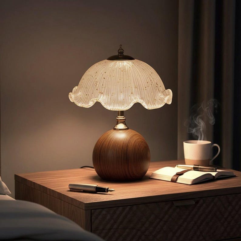 K&ouml;nnte beinhalten: Eine Tischleuchte mit kugelf&ouml;rmigem Holzfu&szlig; und einem geriffelten, durchscheinenden Glasschirm mit goldenen Sprenkeln. Die Lampe steht auf einer Holzoberfl&auml;che neben einem offenen Notizbuch, einem F&uuml;llfederhalter und einer dampfenden Tasse.