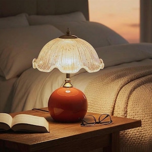 K&ouml;nnte beinhalten: Eine Tischleuchte mit einem gl&auml;nzenden, kugelf&ouml;rmigen orangefarbenen Fu&szlig; und einem durchscheinenden, geriffelten Glasschirm mit dezentem Goldsprenkelmuster. Die Lampe strahlt ein warmes Licht aus und beleuchtet eine Holzfl&auml;che mit einem offenen Buch.