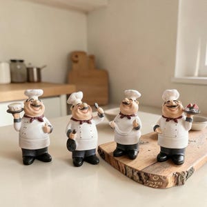 Op de afbeelding: Vier keramische chef-figuren, elk met een wit uniform, een zwarte broek en een hoge witte hoed. De chefs houden verschillende items vast, waaronder een cake, een pan en een lepel. Ze hebben lachende gezichten en rode strikjes. De beeldjes staan op een houten snijplank.