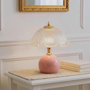 K&ouml;nnte beinhalten: Eine Tischleuchte mit einem runden, massiven rosa Keramikfu&szlig; und einem goldfarbenen Metallhals. Der Lampenschirm ist aus geriffeltem, gewelltem Glas gefertigt, transparent und mit goldenen Sprenkeln im gesamten Glasmaterial versehen.