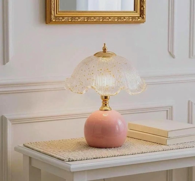 K&ouml;nnte beinhalten: Eine Tischleuchte mit einem runden, massiven rosa Keramikfu&szlig; und einem goldfarbenen Metallhals. Der Lampenschirm ist aus geriffeltem, gewelltem Glas gefertigt, transparent und mit goldenen Sprenkeln im gesamten Glasmaterial versehen.
