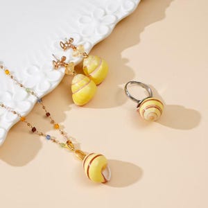 May include: A jewelry set consisting of a necklace, a pair of earrings, and a ring. Each piece features a yellow shell-shaped charm with brown spiral stripes. The necklace has a delicate gold chain with small multicolored beads, and the earrings have gold-tone metal accents.