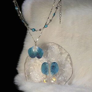 May include: A silver-toned necklace featuring a double chain with small blue crystal accents and a pendant shaped like a blue seashell. The set includes matching drop earrings, each with a blue seashell top and a small, iridescent white bead.