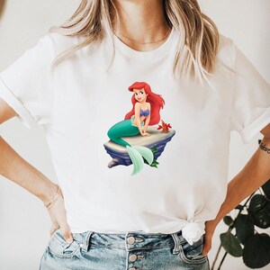 May include: White t-shirt featuring a colorful graphic of a mermaid with red hair, a blue top, and a green tail, sitting on a rock. A red crab is also visible. The shirt is made of a soft material.