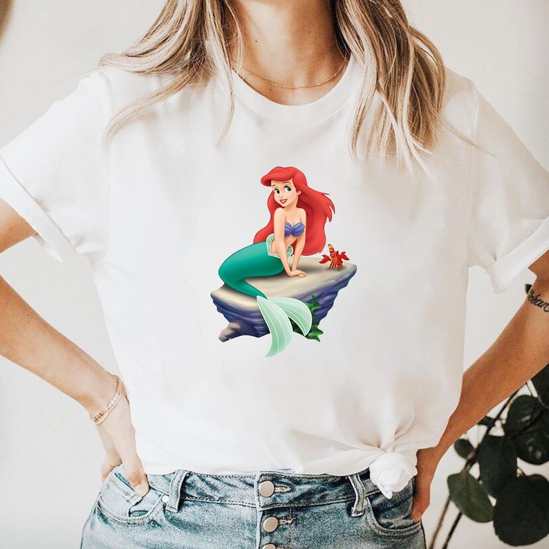 May include: White t-shirt featuring a colorful graphic of a mermaid with red hair, a blue top, and a green tail, sitting on a rock. A red crab is also visible. The shirt is made of a soft material.