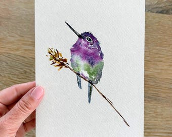 May include: Watercolor painting of a hummingbird perched on a branch. The bird features purple, green, and blue hues, with a long black beak. The artwork is on a white rectangular paper, with a wooden background.