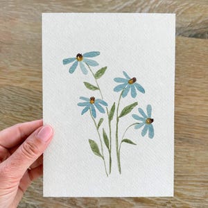 May include: A rectangular piece of textured white paper featuring a watercolor painting of four light blue flowers with dark brown centers and green stems with leaves. The artwork is centered on the paper, showcasing a simple botanical illustration.