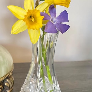 May include: A cut-crystal vase displays a small floral arrangement. The flowers are yellow daffodils and a single purple flower. The vase is on a dark wooden surface. The vase has a cut-glass design.