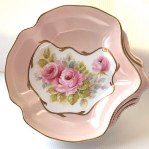 May include: A pink porcelain serving dish with a scalloped edge and gold trim. The center features a floral design of pink roses and green leaves on a white background. The dish has a wavy, irregular shape.
