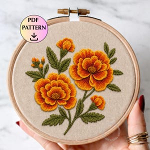 May include: An embroidery hoop featuring a detailed floral design with orange and yellow marigold flowers and green leaves. The text "PDF PATTERN" is displayed on the top left. The embroidery is set against a beige fabric.