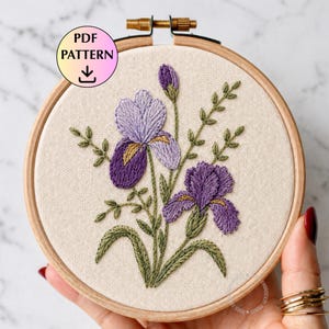 Puede incluir: Un bastidor de bordado con un diseño floral detallado de iris moradas con hojas y tallos verdes. El bordado está sobre un fondo de tela beige. El texto "PDF PATTERN" se muestra en la parte superior izquierda.