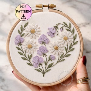 May include: Embroidered floral design in a wooden hoop. The design features white daisies, lavender flowers, and green leaves arranged in a heart shape. The text "PDF PATTERN" is visible in the upper left corner.