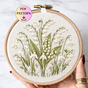 May include: Embroidered floral design of white lily of the valley flowers and green leaves in a wooden embroidery hoop. The text "PDF PATTERN" is in a circle at the top left with a downward arrow.