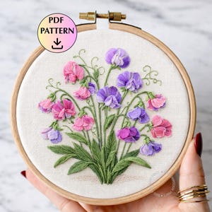 以下が含まれることがあります： ピンクと紫のスイトピーの花柄の手刺繍。クリーム色の生地に刺繍され、木製のフレームで囲まれています。「PDF PATTERN」の文字が円で囲まれています。